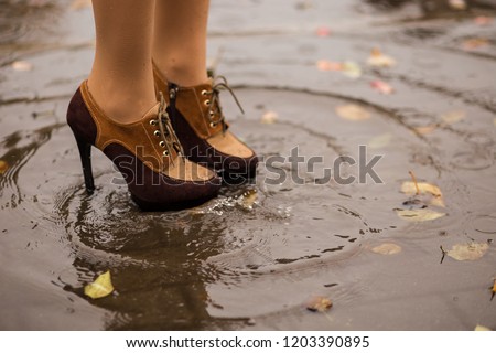 close up of young Caucasian woman feet in brown boots standing in a puddle with leaves and raindrops under rain on a city street