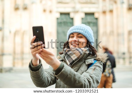 Happy travel woman taking photo with her phone. Autumn or winter trip. Young woman in warm clothes enjoying journey in Europe. People, lifestyle and travel concept. Girl tourist walking on the street
