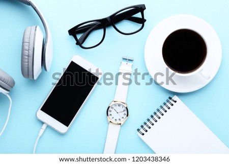 Smartphone with headphones, cup of coffee, notebook and glasses on blue background
