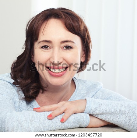 Lovely middle-aged brunette woman with a beaming smile sitting on a sofa at home looking at the camera