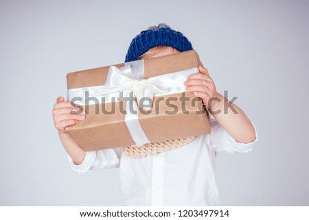 Happy little smiling blonde curly hairstyle boy in a knitted blue hat with christmas gift box with bow on white background in studio. new Year present in the hands of a male child