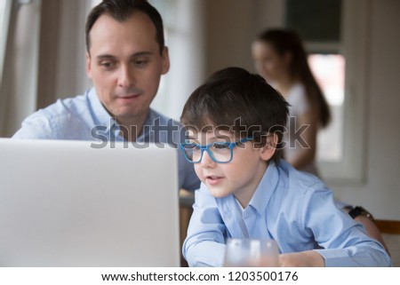 Young handsome man little preschool boy in eyeglasses looking at computer sitting together at home. Father spend free time on weekend with small son using laptop having fun watching recreational video
