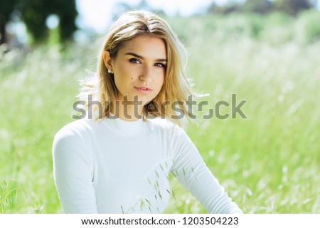 Attractive young blonde Caucasian woman in tight white top and denim posing outside in long grass