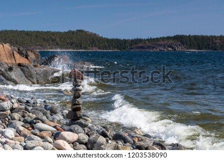 Stacked stones on the seashore and waves in foreground, cliffs and forest and a blue sky in background, picture from High Coast area in Northern Sweden.