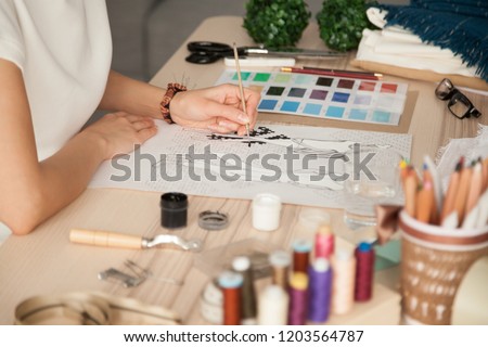 Female clothing designer sketching, drawing with brush at workplace close up in cozy workshop studio, fashion designer, dressmaker creating new fashionable handmade clothes collection, hands view