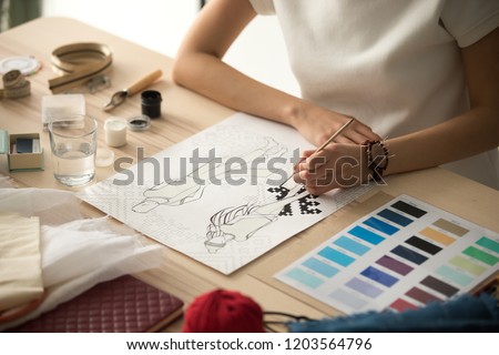 Female clothing designer coloring sketch, drawing with brush at workplace close up in workshop studio, fashion designer, dressmaker creating new fashionable handmade clothes collection, hands view