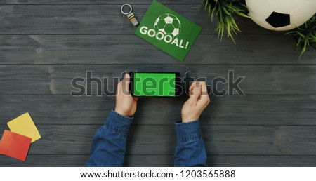 Top view on the black horizontal smartphone with green screen in hands of man who scrolling and taping on the dark wooden table with football ball and writing Goal. Chroma key.