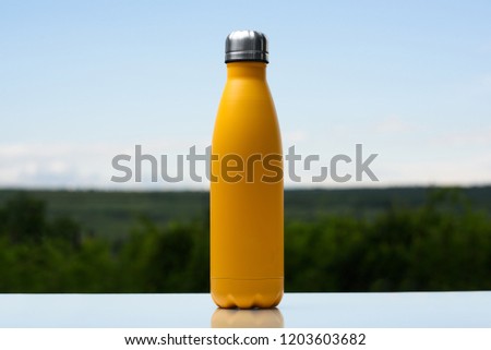 Thermo stainless bottle for water, tea or coffee. Sky and forest on background. On the glass desk. Thermos of matte yellow color.