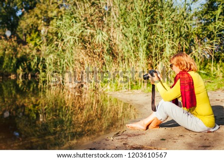 Middle-aged woman checking images on camerasitting by autumn river bank. Senior woman enjoying nature and hobby shooting photos