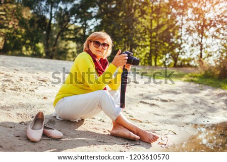 Middle-aged woman checking images on camerasitting by autumn river bank. Senior woman enjoying nature and hobby shooting photos