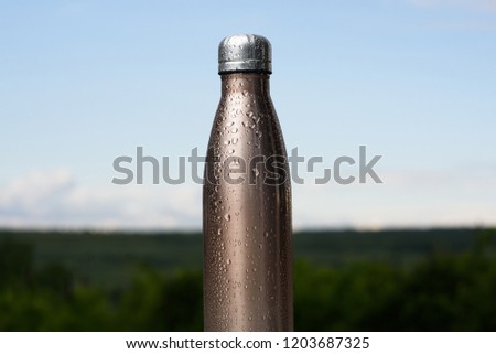 Thermo stainless bottle, sprayed with water. Sky and forest on background. Thermos of maro color.