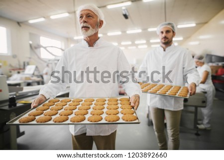 Picture of two male food factory employees in sterile clothes holding trays full with fresh cookies. Working on production line in food factory.