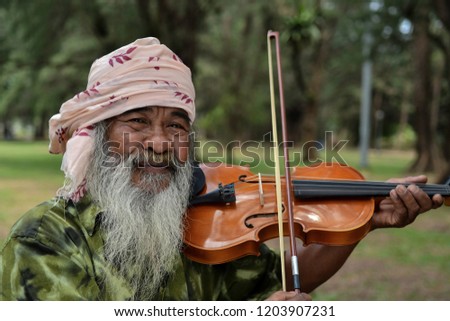 Horizontal photo of Senior Asian man playing violin outdoors with bright green trees in background