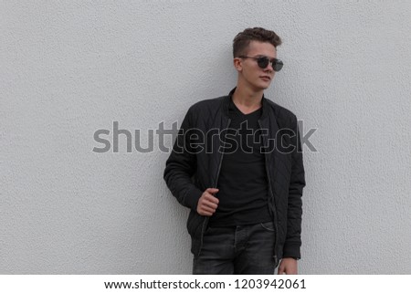 Fashionable young guy with sunglasses in a black jacket and a black t-shirt near the gray wall