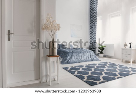 Flower in silver vase on the wooden table next to closed door to elegant new york style bedroom with patterned carpet and white furniture, real photo