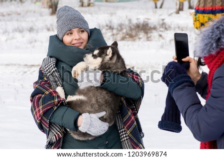 Waist up portrait of modern young woman holding cute Husky puppy and looking at smartphone camera with friend taking photo of her outdoors in winter