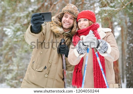 Waist up portrait of active young couple taking selfie photo via smartphone while enjoying skiing in snowy winter forest during date