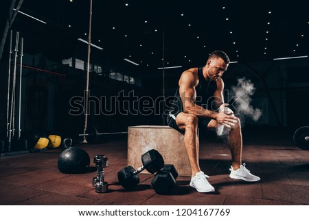 handsome athletic man sitting on cube with gym equipment around and clapping hands with talc before workout Royalty-Free Stock Photo #1204167769