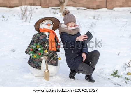 Adorable young girl is taking pictures of selfie with a snowman in beautiful winter park. Winter activities for children. Close up