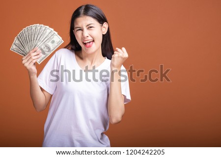 Woman holding cash notes isolated in orange background. Young asian woman in white t-shirt in winning pose, holding dollar note. Young rich hipster concept. Royalty-Free Stock Photo #1204242205