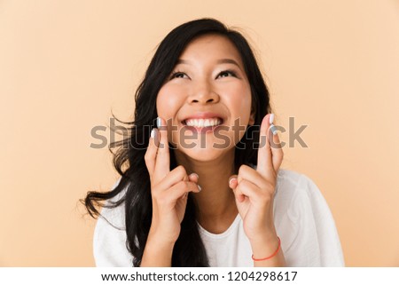 Portrait of a smiling young asian woman isolated over beige background, holding fingers crossed for good luck