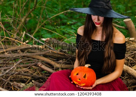 Halloween holiday background. Halloween Witch with a halloween pumpkin jack o lantern decor with funny face in a dark forest. Beautiful young woman in witches hat and costume holding pumpkin.