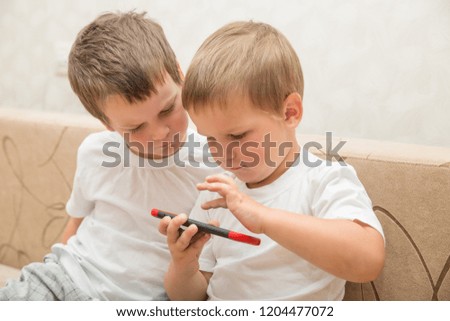 Two little boys in white T-shirts are sitting on sofa at home. One boy plays games on smartphone,  second one looks