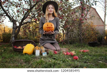 Beautiful women in black hat holding Jack o'lantern and sitting in the garden . pumpkins, candle and apple around. Halloween concept 