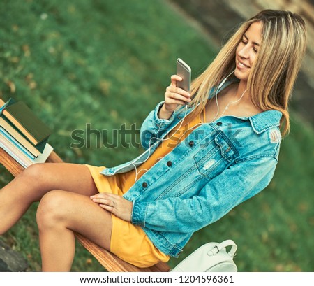 Many books are on a wooden bench. Next to the books is a student in a yellow dress and a denim jacket. She looks into the phone and listens to music through the headphones.