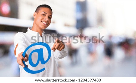 Young african american man with white shirt holding icon of at dot com and pointing to the front in the middle of the city
