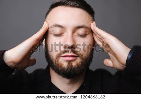 Handsome bearded male keeping eyes closed while standing on gray background and thinking