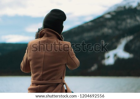 Girl in black toque looks towards the rocky mountains in Canmore, Alberta - she wears a stylish brown coat lined with fur 