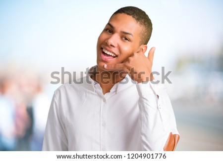 Young african american man with white shirt making phone gesture and speaking with someone. Call me back sign on unfocused outdoor background