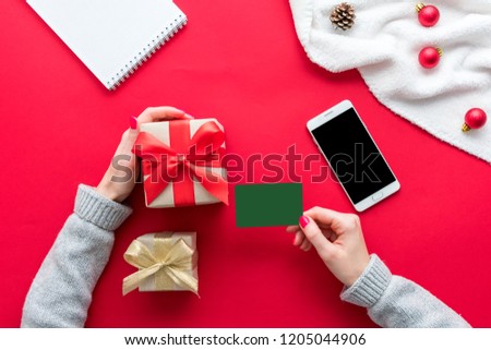Female hands, woman holding a business card and gift. White smart phone and Notepad on the red table, gifts boxes for Christmas and New year, background with copy space, for advertisement, top view