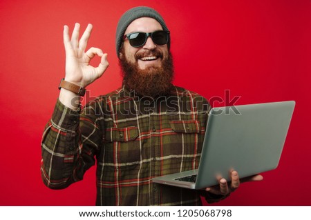Cheerful bearded hipster man wearing sunglasses and holding laptop and showing OK gesture
