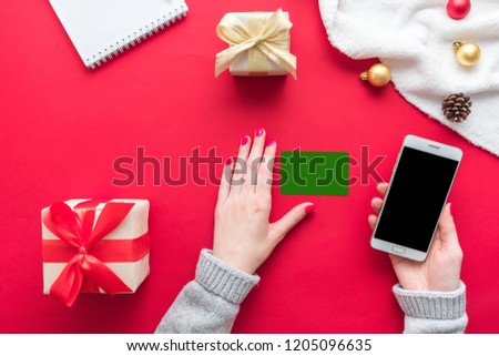 Female hands, woman holding a white smart phone, business card and Notepad on the red table, gifts boxes for Christmas and New year, background with copy space, for advertisement, top view