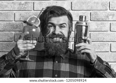 Barman with beard, stylish hair and cheerful face on light brick wall background. Lets party concept. Hipster prepared cocktail. Man holds glass, delicious cocktail with orange, straw and shaker.