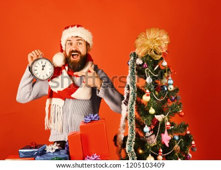 Guy in red hat with xmas gift box points at alarm clock. Man with beard and surprised face on red background. Santa with red presents near decorated fir tree. Christmas gifts and time concept