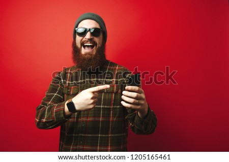 Cheerful amazed bearded hipster man pointing at phone over red background