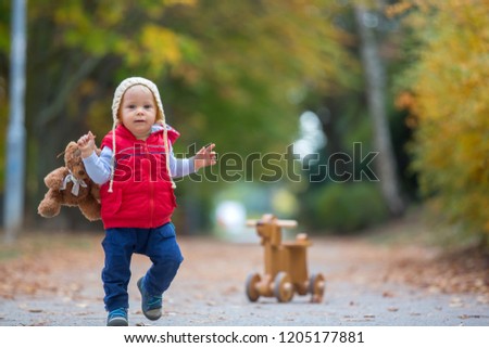 Little toddler boy with teddy bear, riding wooden dog balance bike in autumn park on a sunny warm day, children leisure activities and happiness concept