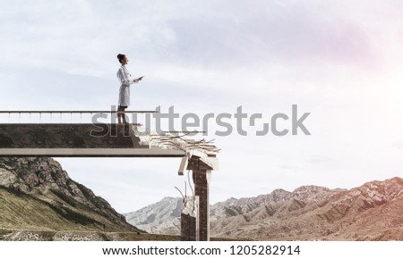 Young but professional female doctor holding tablet in hands while standing at the end of broken bridge with landscape view on background. Medical industry concept