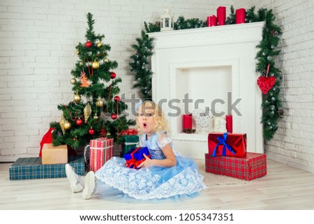 Cute little girl in a nice dress with long curly blondу hair holding a box with a gift at home near a Christmas tree with gifts and garlands and a decorated fireplace .new Year morning