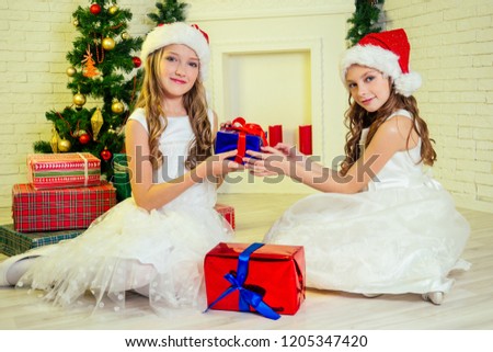 Cute little twin girls in Santa Claus hat curly blondу hair opening Xmas presents at home near a Christmas tree with gifts, garlands and a decorated fireplace .Family on Christmas eve new Year morning
