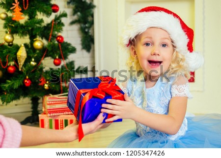 Mother holding on hand Christmas gift box for daughter little girl in Santa Claus hat curly blondу hair at home near a Christmas tree with gifts, garlands and a decorated fireplace .new Year morning