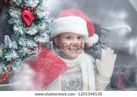little girl in santa hat and with gifts is looking into the snow-covered window from a winter street
