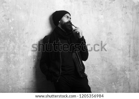 Biker fashion and beauty. Guy with serious face. Man with long beard and mustache. Fashion model on textured wall background. Hipster in leather jacket and hat with glasses.