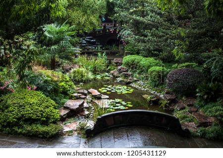 Stone Bridges in a Chinese Garden. Traditional Garden Design with Stone Bridge over a Small Pond with lotus leaves inside. Surrounded by Trees and Flowers on a Rainy Day.
