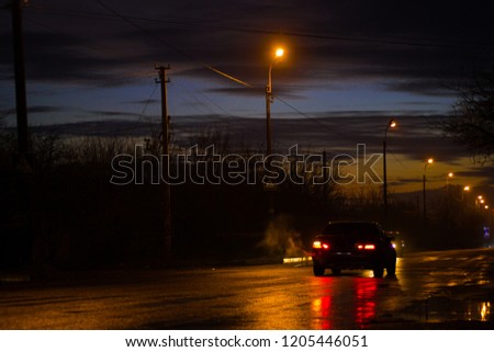 A black car is standing on the road waiting to turn left with an oncoming car. Evening autumn motive, night road in a long evening. From the exhaust pipe of the car is light exhaust smoke.