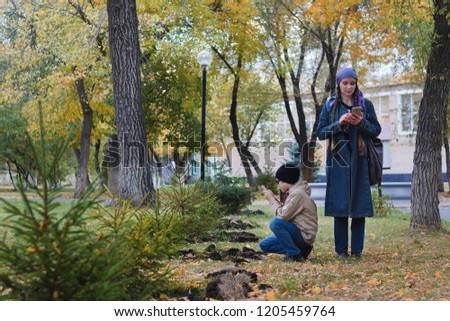 A woman and her child taking pictures on a mobile phone of a small tree in the Park in the fall.