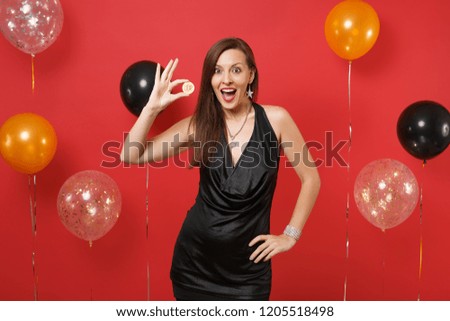 Excited young woman in black dress celebrating holding bitcoin metal coin of golden color, future currency on bright red background air balloons. Happy New Year, birthday mockup holiday party concept
